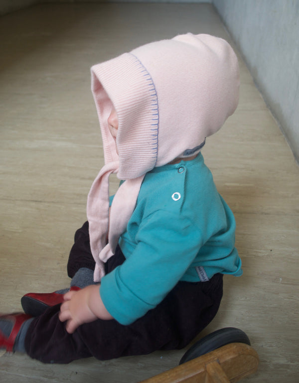 Child wearing a kin and Cloth soft pink hooded cashmere handcrafted luxury bonnet  with hand detailed embroidery sitting down on the floor.